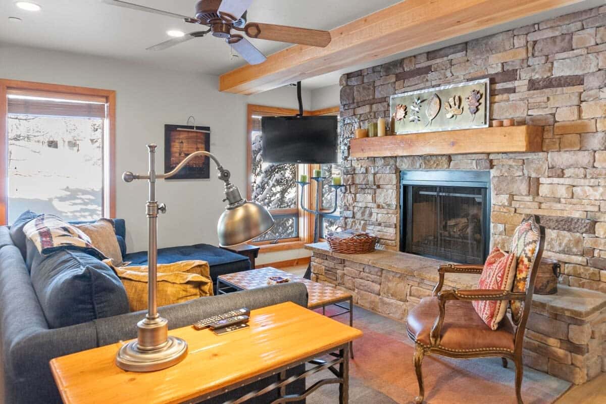 The first living room features a large sectional sofa with a chaise on the right side, a leather chair, a mounted 37” flat-screen TV, and a stone hearth double-sided fireplace.