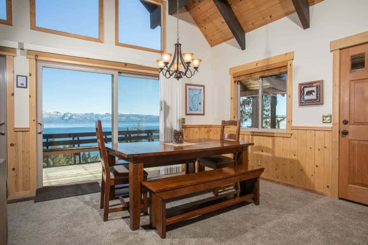 The dining area features a wooden dining table with chairs and bench seating for 8 next to the floor-to-ceiling windows overlooking the lake providing a stunning view to enjoy while you eat.