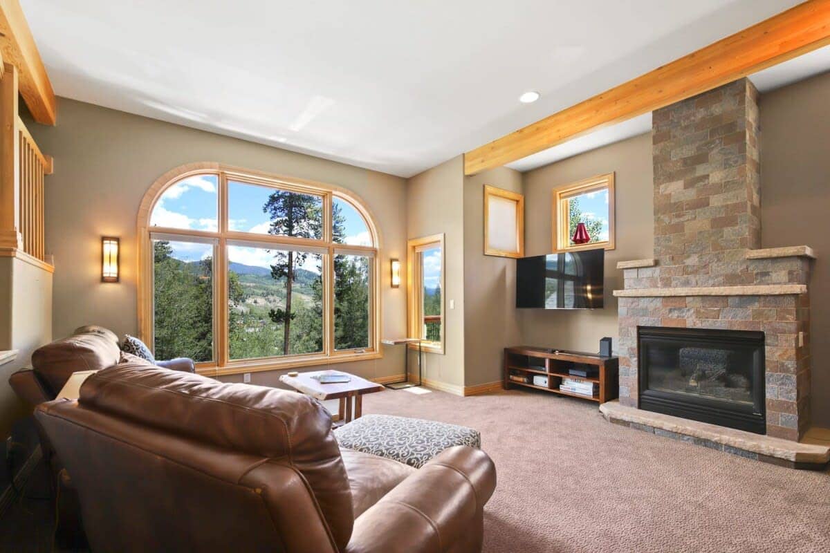 The spacious living room features wood beam accents, abundant natural light, two leather couches, a roomy leather armchair with an ottoman, coffee table, end table, lamps, a flat-screen TV and a striking, stone fireplace.