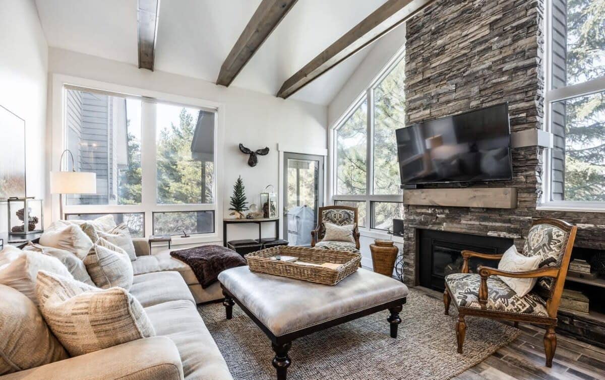The living room in this home has it all - a roomy sectional sofa, oversized coffee table/ottoman, two reading chairs, a large flat-screen TV, gas fireplace with stone work, natural light, tree views and stylish wood beams.